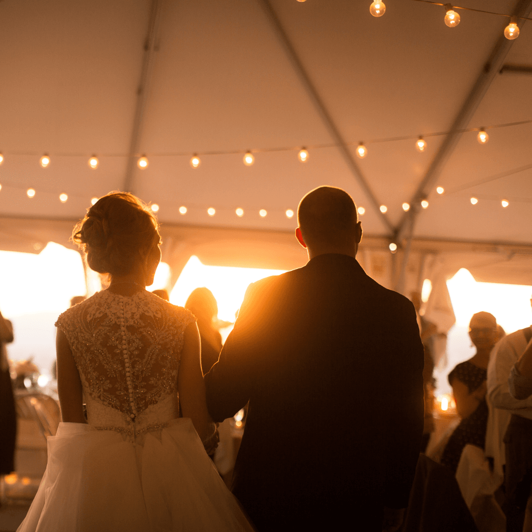 A bride and groom stand together in soft golden light under a tent adorned with string lights.