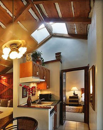 A compact, brightly lit kitchenette in an attic space with a sloped wood-beam ceiling and skylight. An open doorway leads to a bedroom.