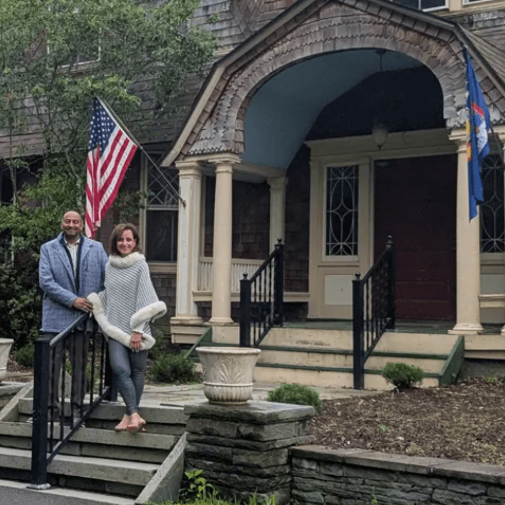 A man and woman pose on the steps of a large, historic home with an American flag in the background.