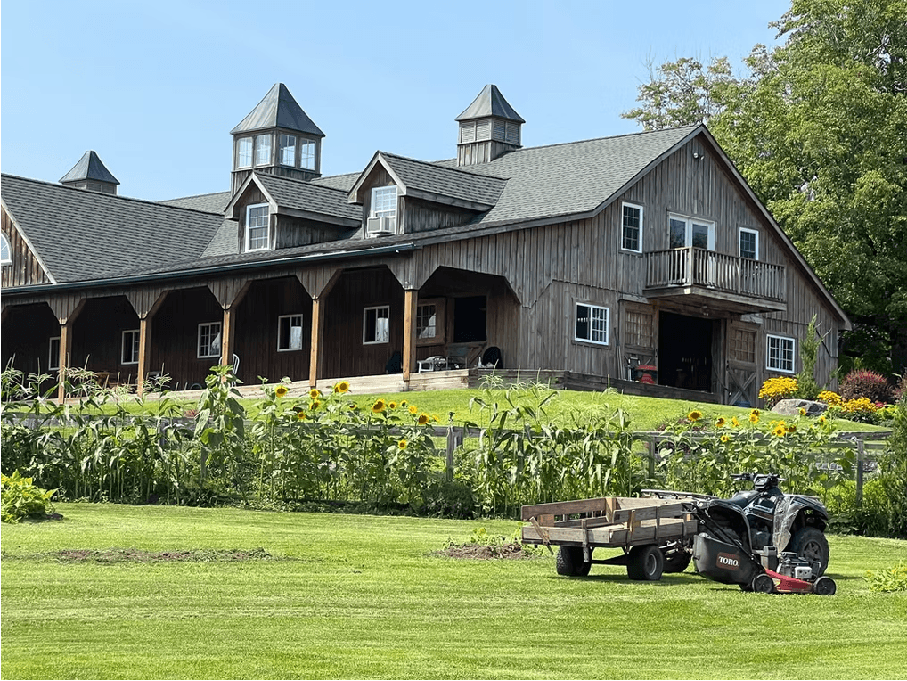 A wooden barn with three cupolas is surrounded by sunflowers and a lawn, featuring a lawn mower and trailer in the foreground.