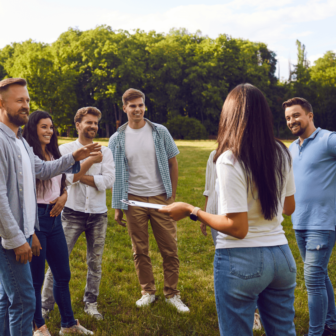 A group of six people socialize in a sunny park, engaging attentively with one person holding a clipboard.