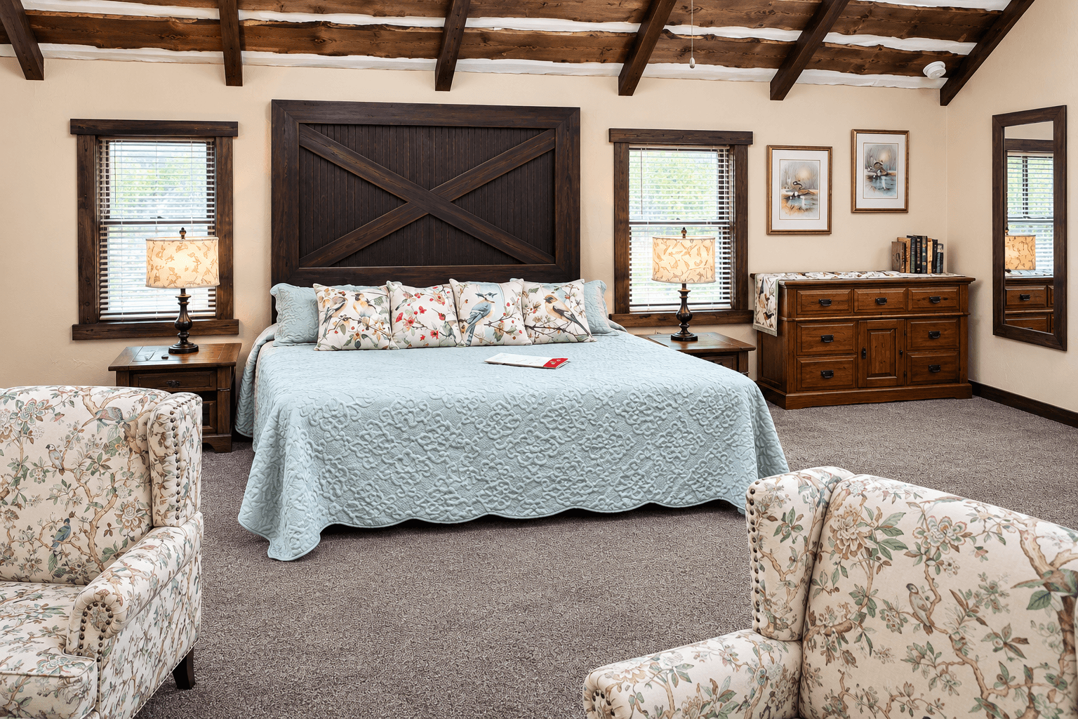 Cozy bedroom featuring a large bed with a light blue quilt and floral pillows, accompanied by patterned armchairs and a wooden dresser.