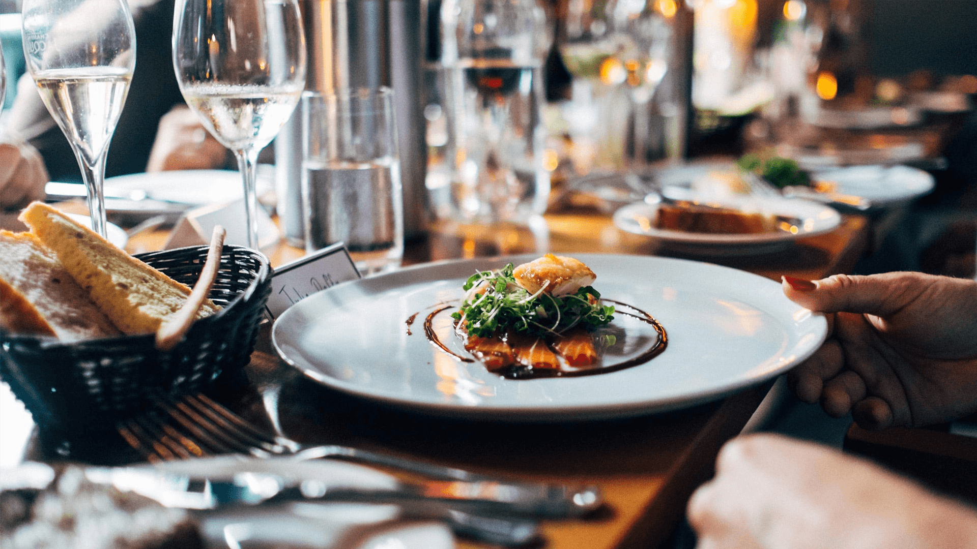 Elegant dining scene with a focus on a gourmet dish topped with greens, surrounded by wine glasses. The ambiance is warm and sophisticated.