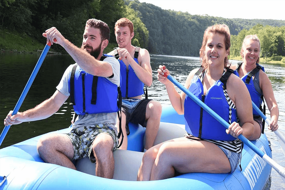 Four people paddle together on a blue raft in a serene river setting.