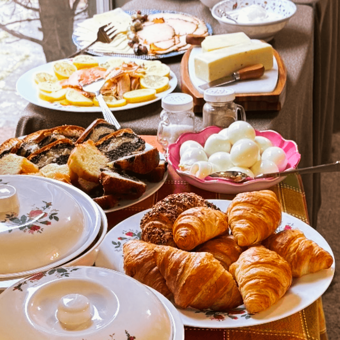 A variety of breakfast foods including croissants, bread, cheese, boiled eggs, and fruit are displayed on a table.