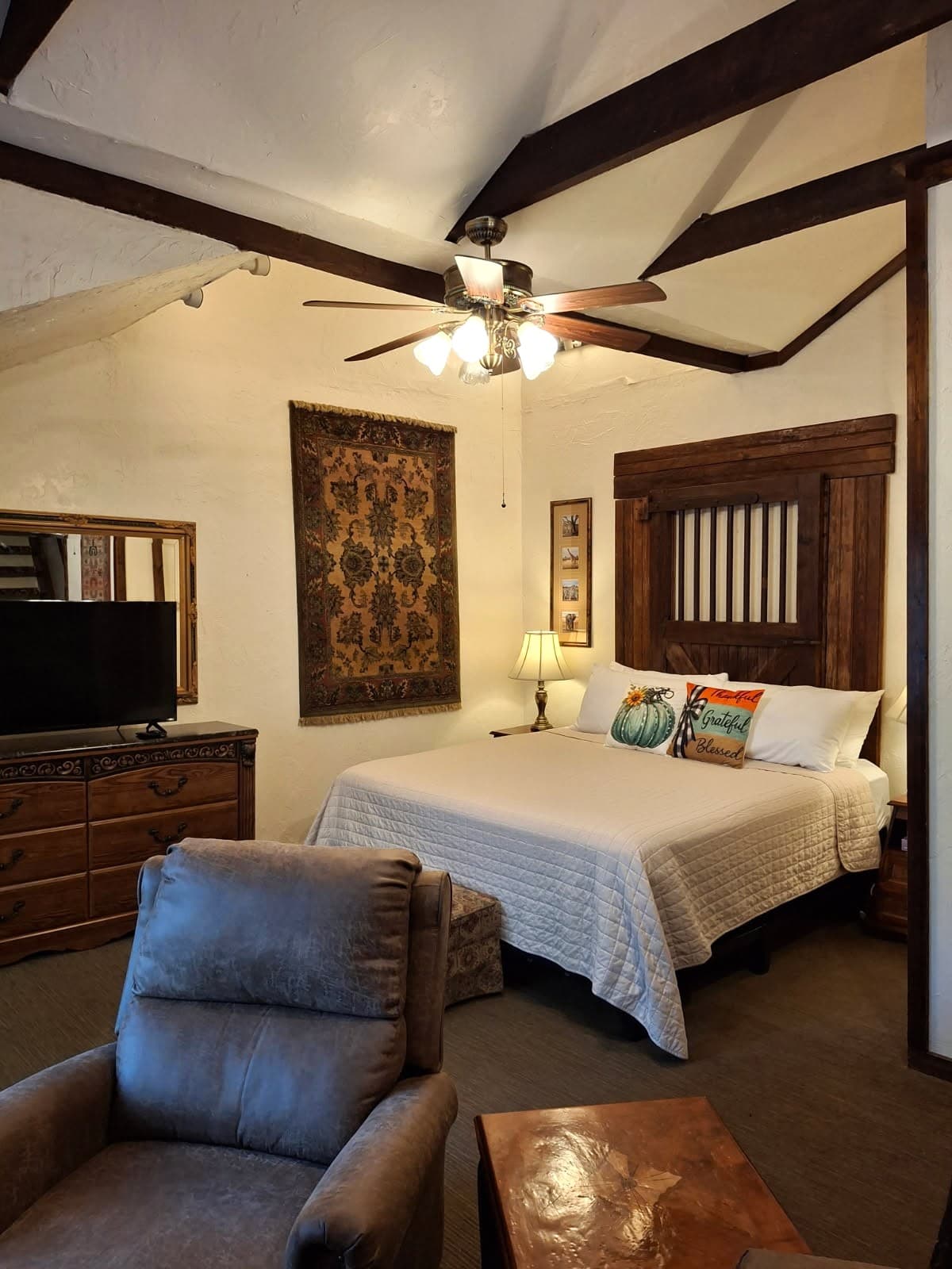 Cozy bedroom with a wooden bed, decorative pillows, a TV, and a reclining chair under a ceiling fan.