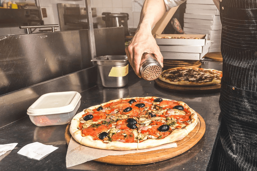 A chef in a kitchen sprinkles seasoning on a freshly made pizza topped with olives, tomatoes, and herbs. The scene conveys a sense of craft and flavor.