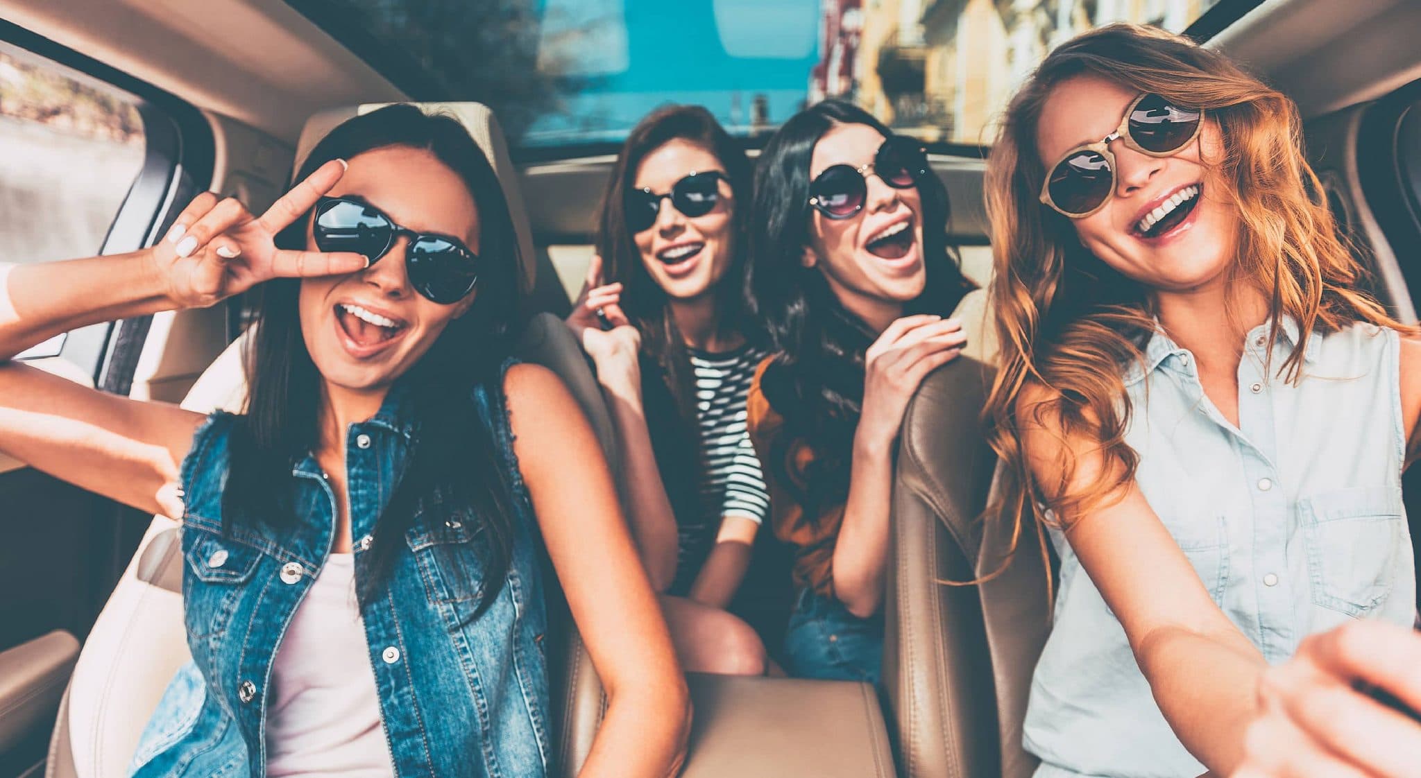 Four young women wearing sunglasses smile and pose for a selfie inside a car.