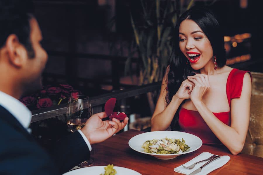 A surprised woman in a red dress reacts joyfully as a man presents her with a ring during a romantic dinner.
