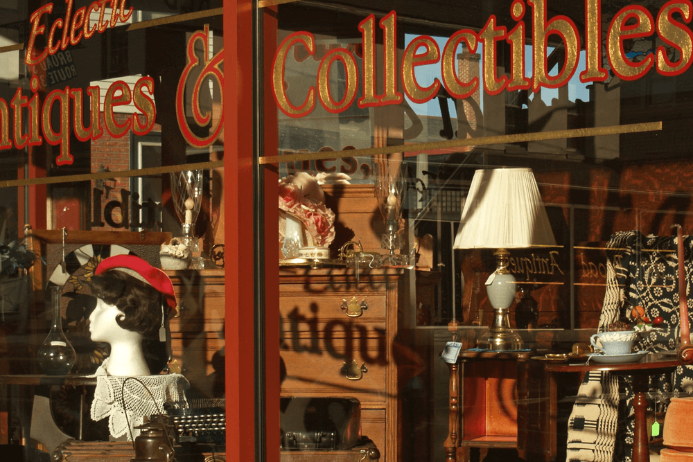 A window display of an antique and collectibles shop featuring vintage items like furniture, a mannequin, and a lamp.