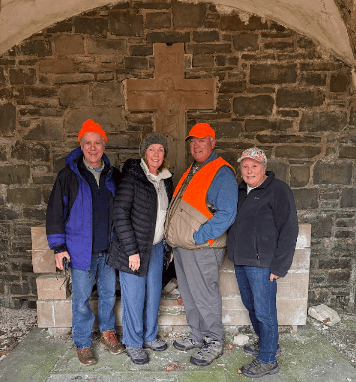Four people in outdoor attire with bright orange hats stand together in a stone shelter featuring a cross in the background.