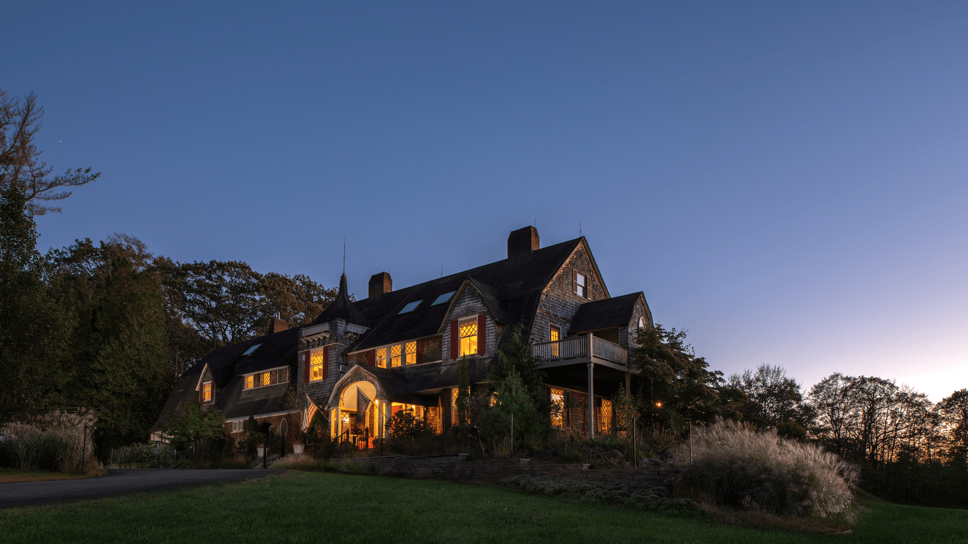 Large, rustic house with warm glowing windows at dusk. Surrounded by trees and grass under a clear, deep blue sky, creating a peaceful ambiance.