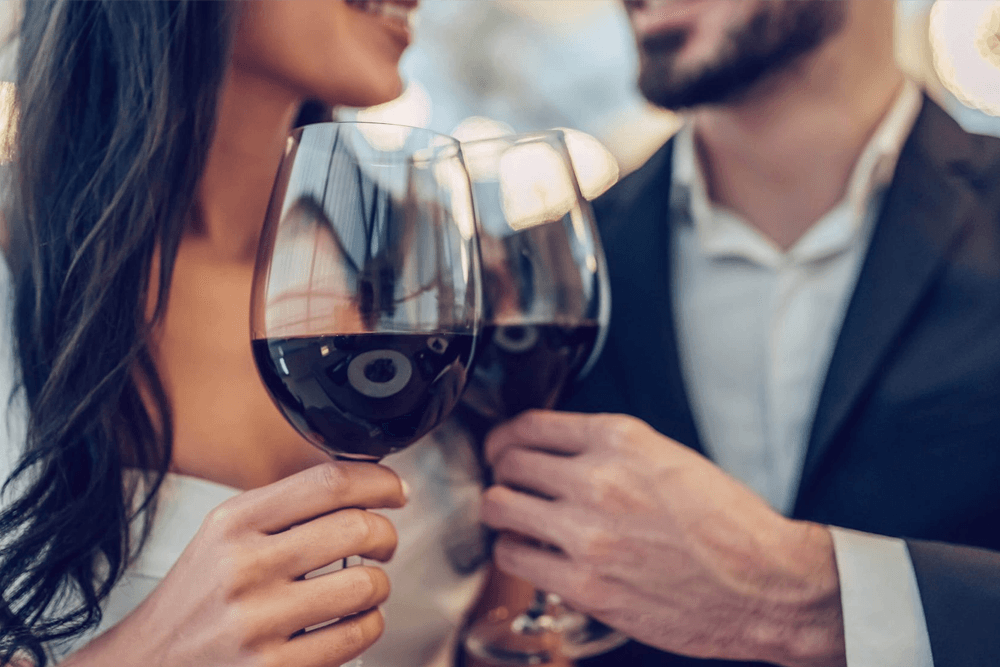 A couple clinks glasses of red wine together, smiling at each other in a romantic setting.