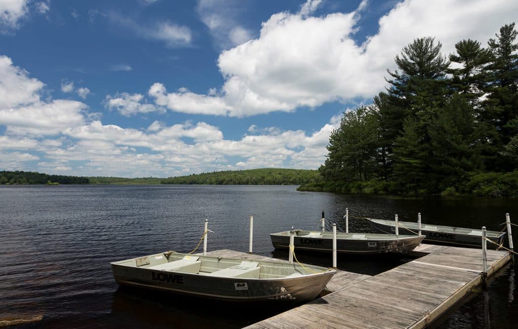 A calm lake with three boats docked at a wooden pier under a partly cloudy sky.