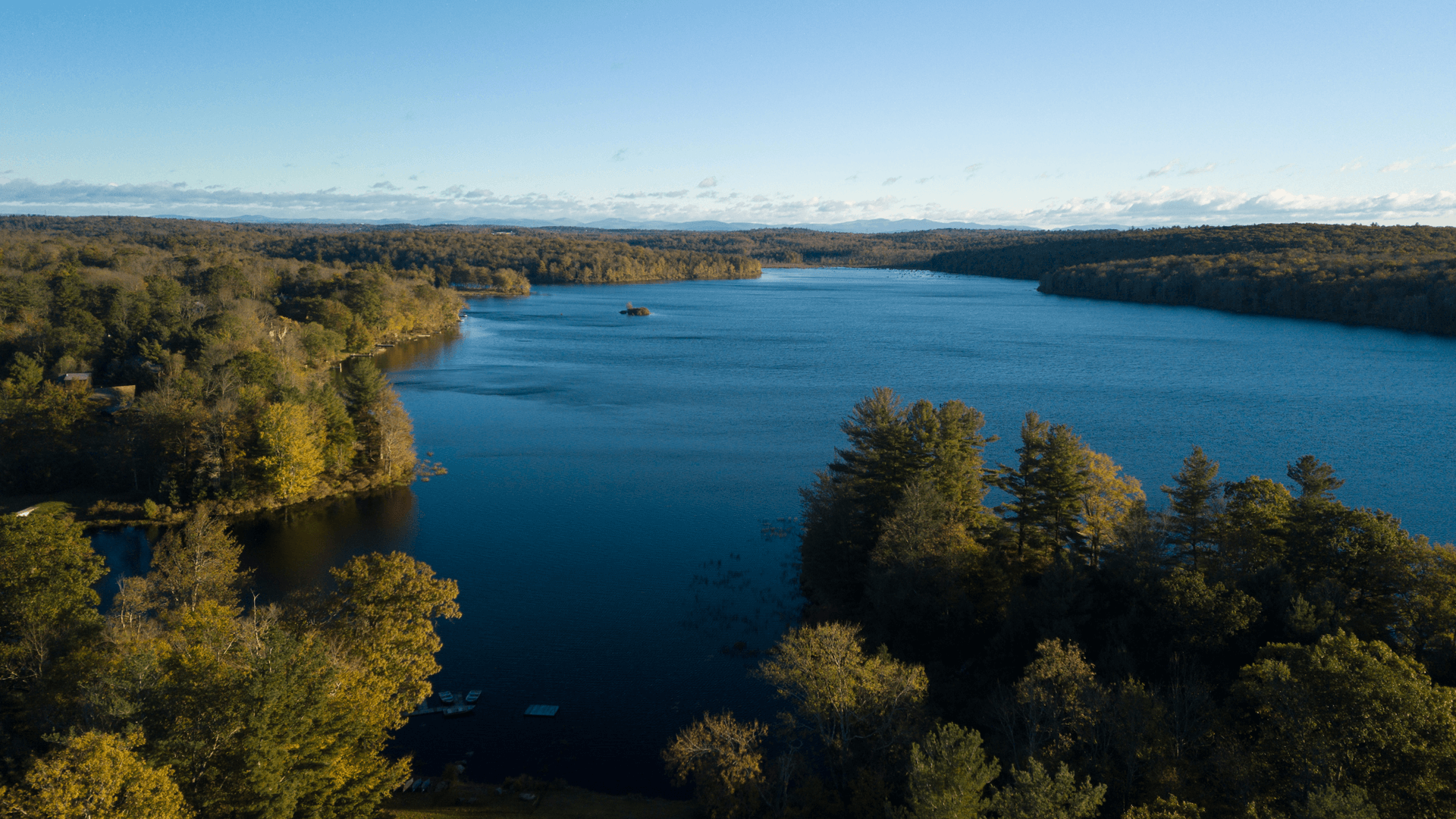 Aerial view of a serene lake surrounded by trees and autumn colors under a clear blue sky.