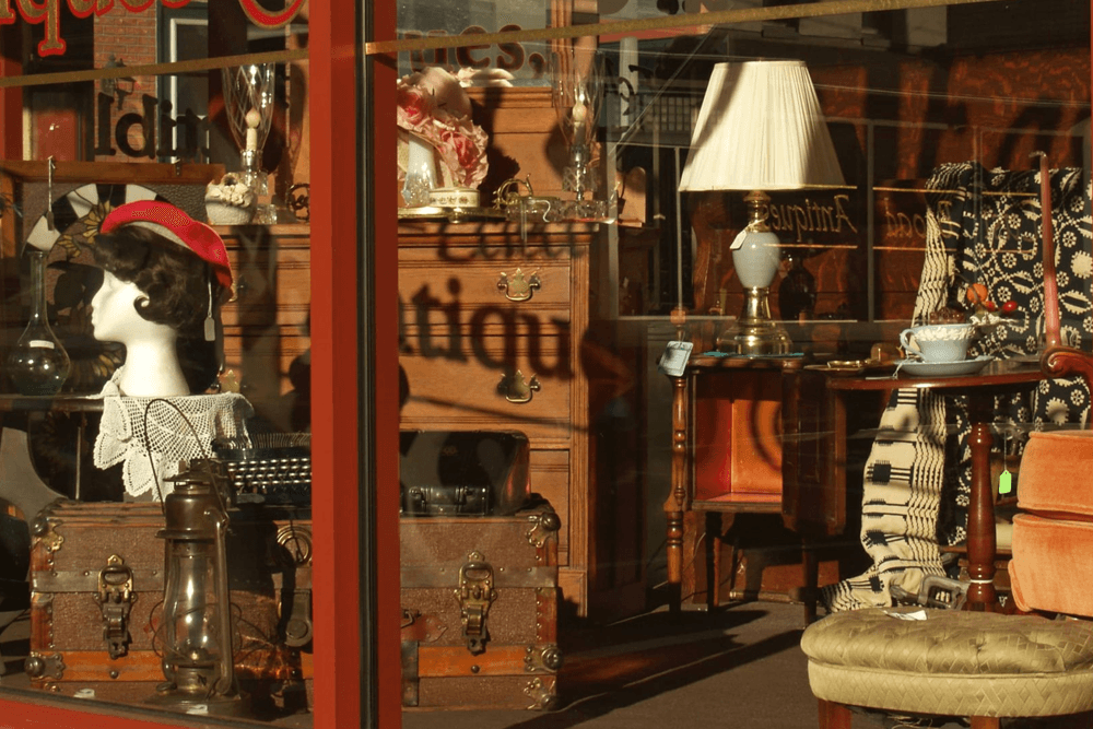 A storefront display filled with vintage furniture and antiques, including a mannequin, lamps, and a typewriter.
