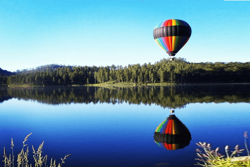 A colorful hot air balloon reflects in a serene lake surrounded by trees.