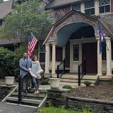 A couple stands together on the steps of a house with an American flag in the yard.