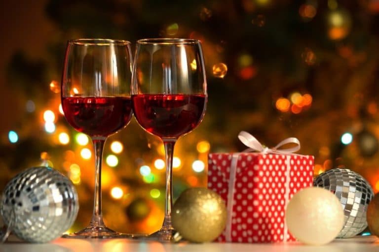 Two glasses of red wine beside a festive gift and decorations, with a blurred Christmas tree backdrop.