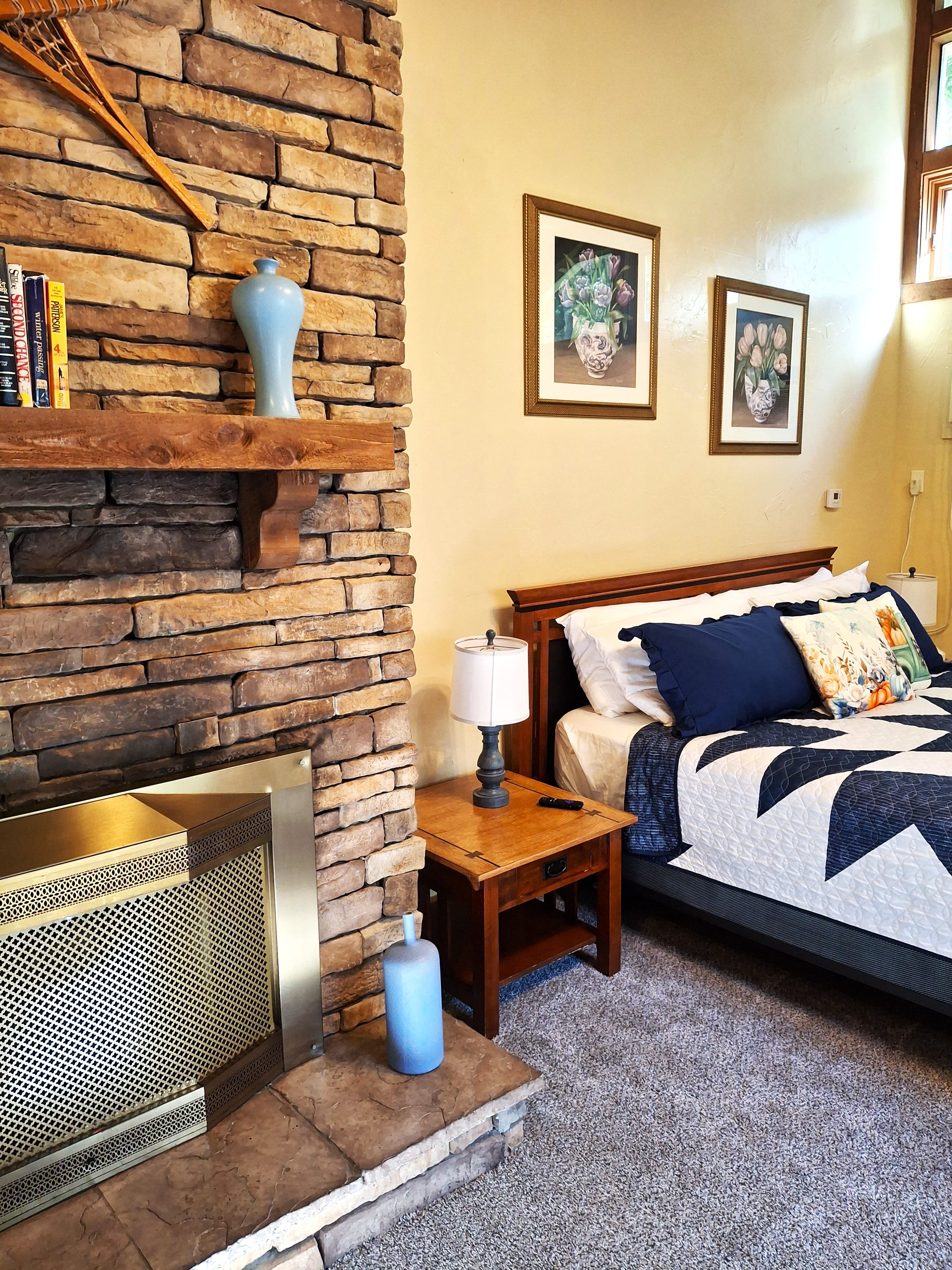 A cozy bedroom featuring a stone fireplace, wooden bed frame, and decorative pillows.