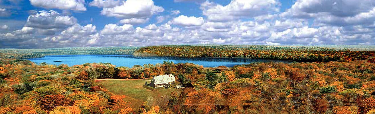 A scenic view of a house surrounded by colorful autumn foliage and a calm lake under a blue sky with clouds.