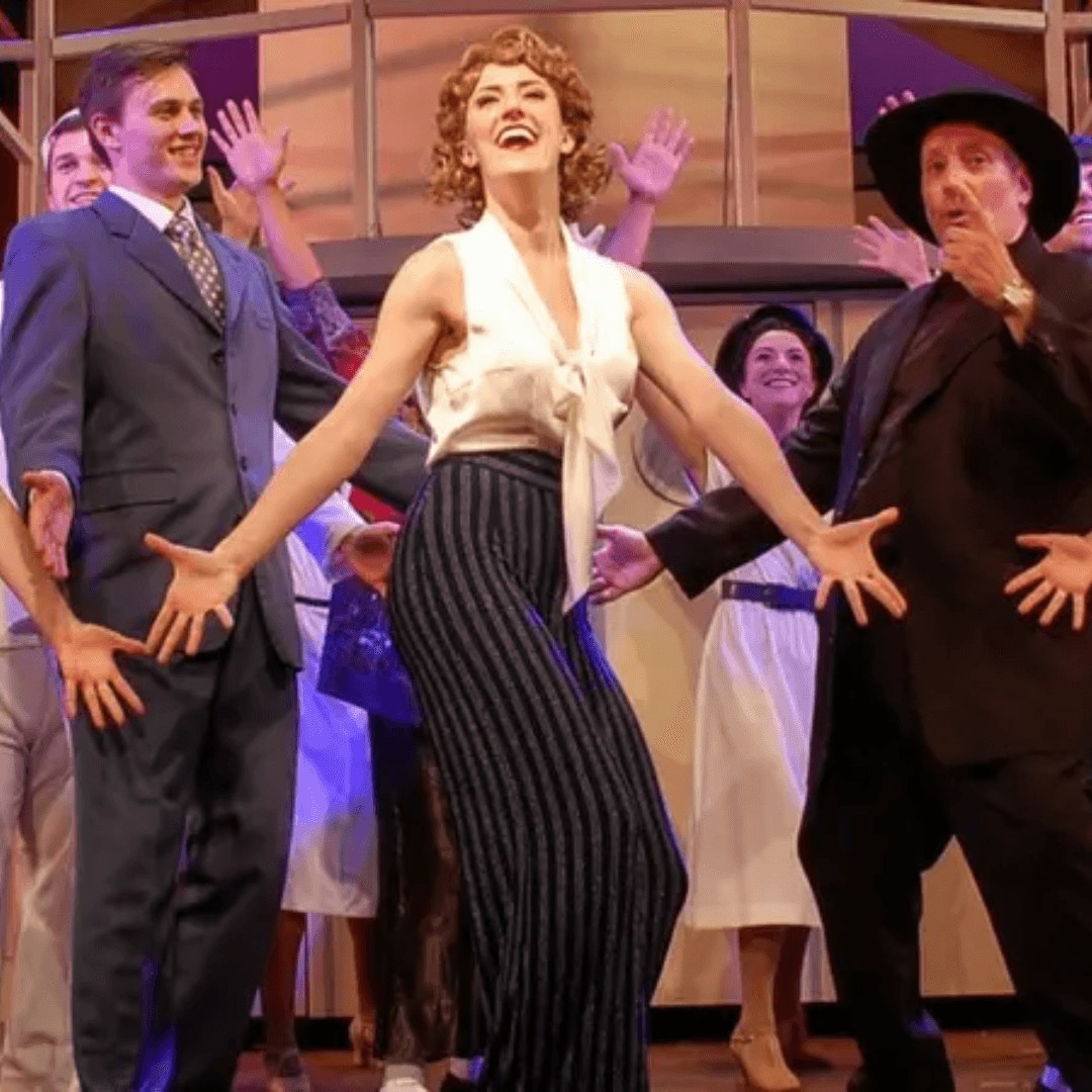 A performer in a white blouse and striped pants joyfully leads a group of cast members on stage.