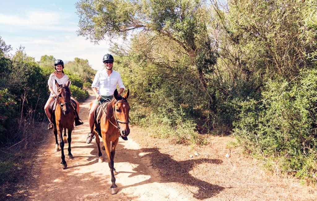 Two riders on horseback along a dirt path surrounded by greenery.