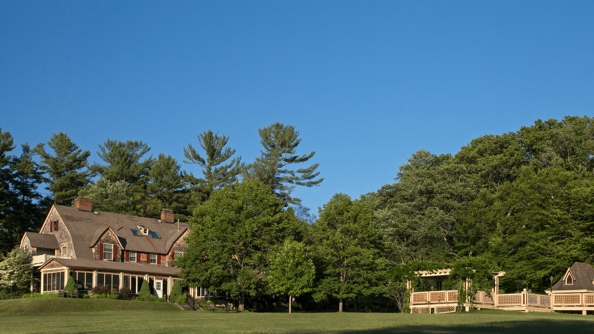 A large house surrounded by lush trees under a clear blue sky. The expansive green lawn conveys a peaceful, idyllic countryside setting.
