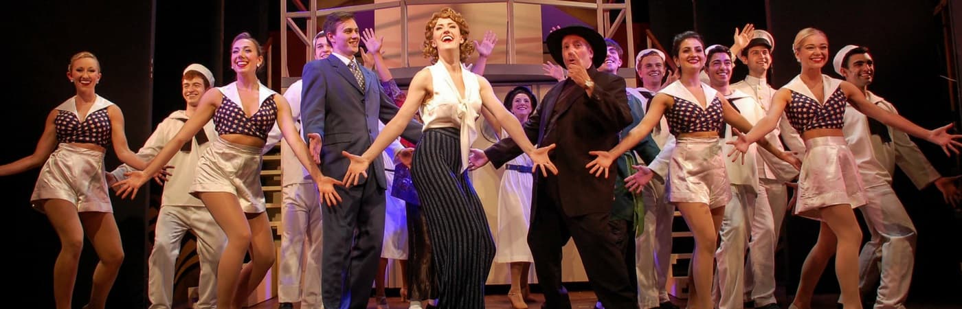 A lively theater performance featuring a large cast in colorful costumes posing and dancing on stage.