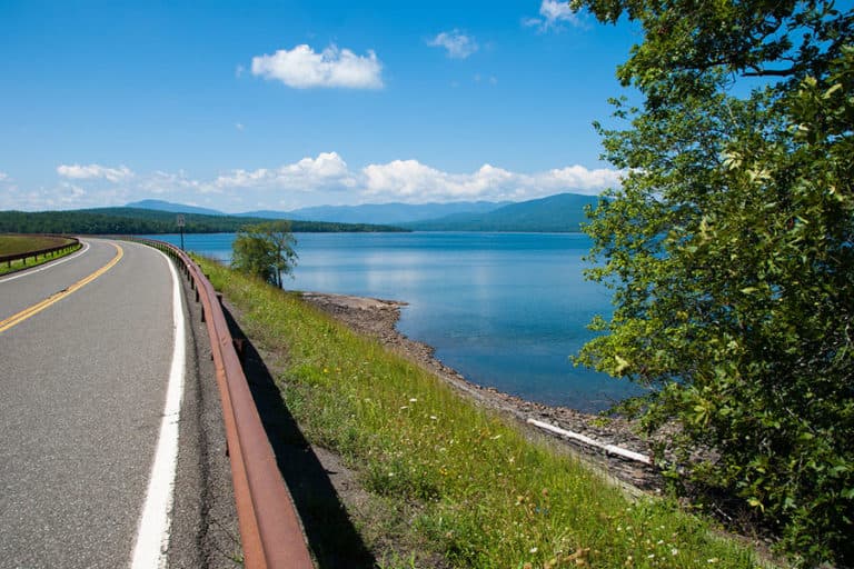 A winding road runs alongside a tranquil lake bordered by greenery and mountains in the distance under a clear blue sky.