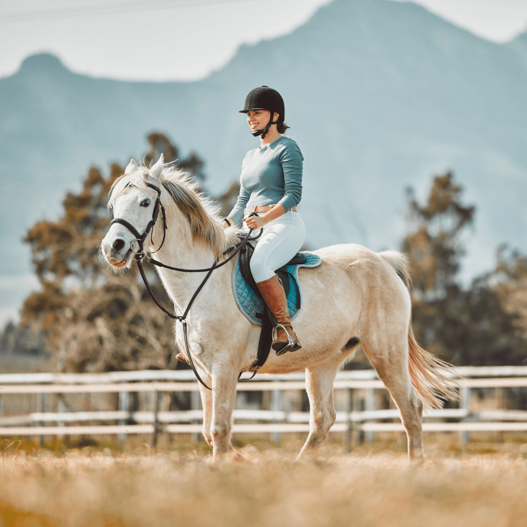 A woman in riding attire smiles while seated on a light-colored horse in a scenic outdoor setting.