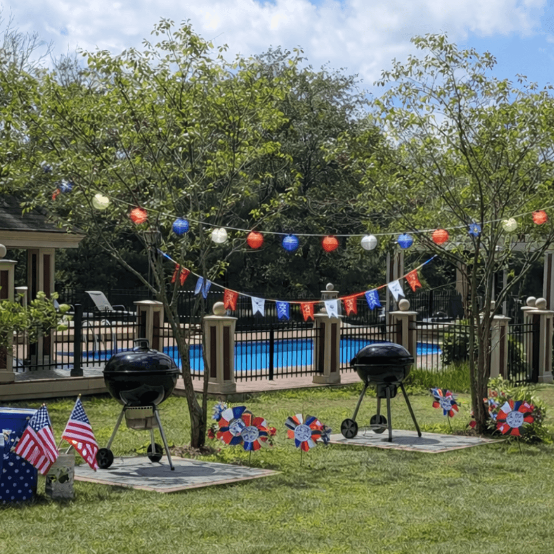 A festive outdoor space featuring two black grills, patriotic decorations, and a swimming pool in the background.