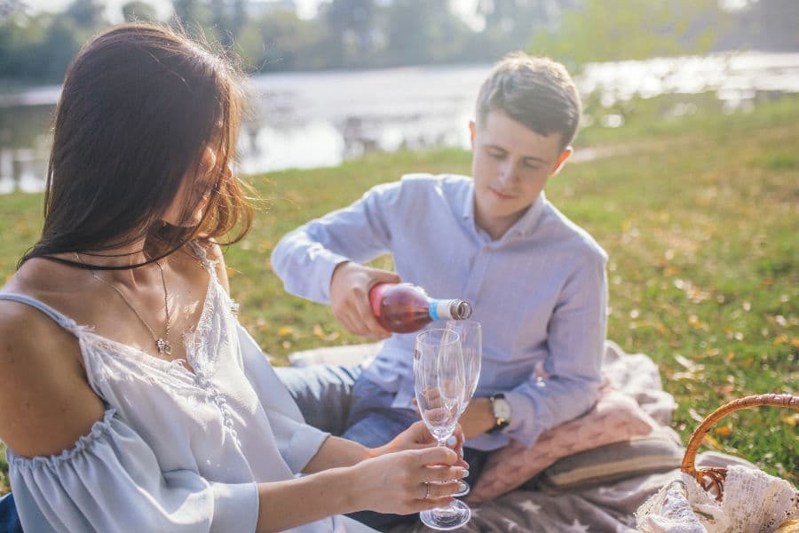 A couple enjoys a picnic by the water, with one person pouring wine into a glass.